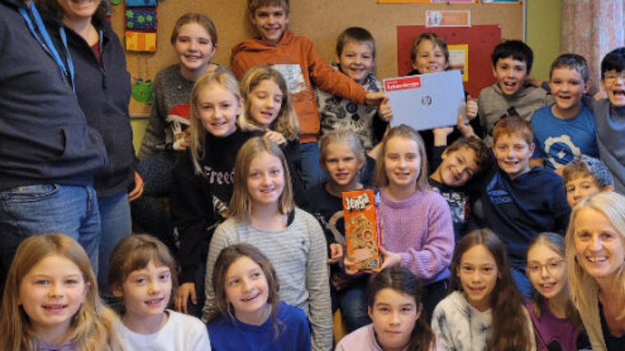 Gruppenfoto in einem Klassenzimmer: Kinder und Erwachsene halten ein „Danke“-Plakat und eine Mappe mit Logo.