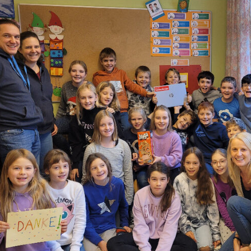 Gruppenfoto in einem Klassenzimmer: Kinder und Erwachsene halten ein „Danke“-Plakat und eine Mappe mit Logo.
