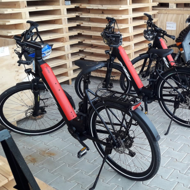 Drei E-Bikes mit roten Akkus stehen vor gestapelten Holzpaletten in einem Lagerbereich.