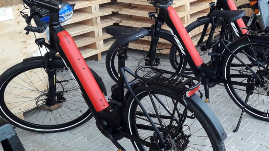 Drei E-Bikes mit roten Akkus stehen vor gestapelten Holzpaletten in einem Lagerbereich.