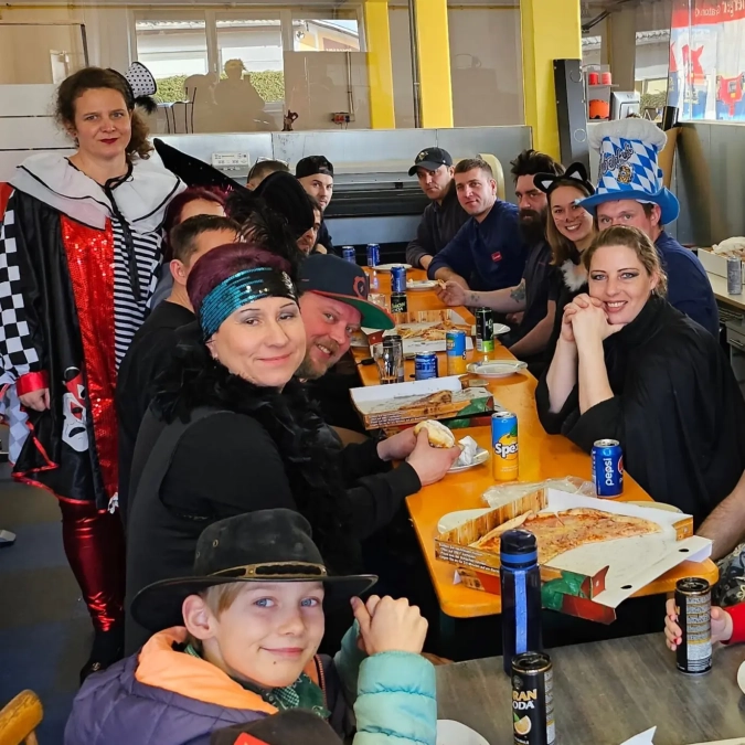 Gruppe in Faschingskostümen sitzt an einem Tisch mit Essen und Getränken in Pausenraum-Umgebung.