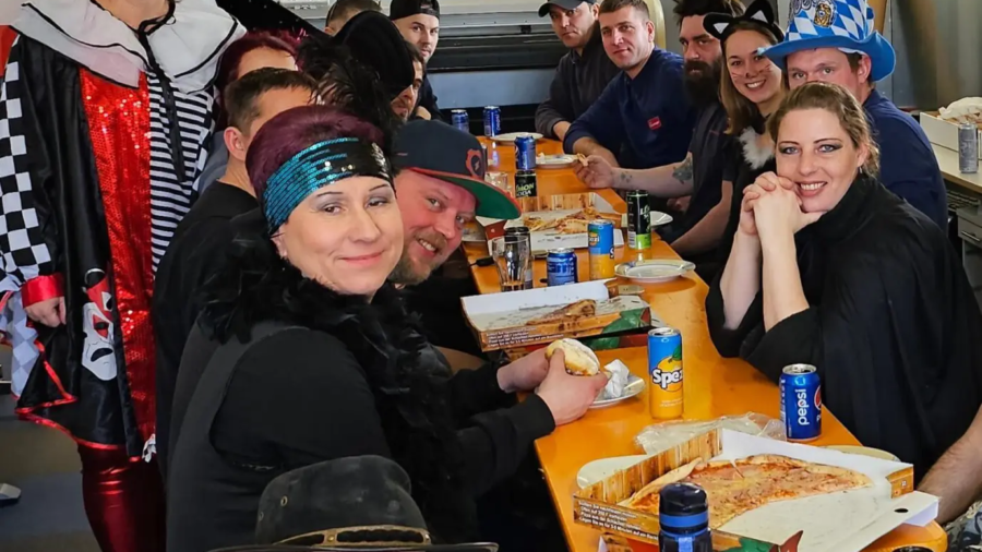 Gruppe in Faschingskostümen sitzt an einem Tisch mit Essen und Getränken in Pausenraum-Umgebung.