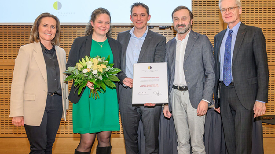 Gruppenfoto bei einer Preisverleihung: mehrere Personen halten Blumen und eine gerahmte Urkunde auf einer Bühne.