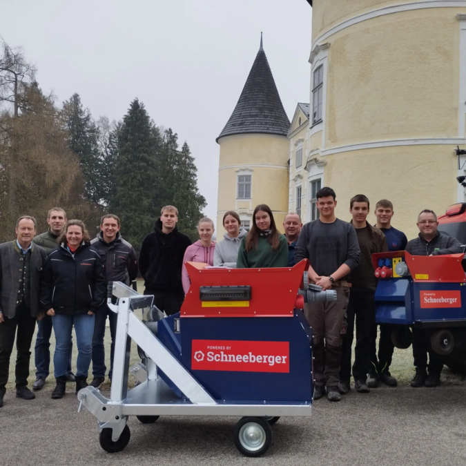Gruppenfoto im Freien: Personen stehen hinter einem rot-blauen Schneeberger Gerät vor einem Gebäude mit Turm.