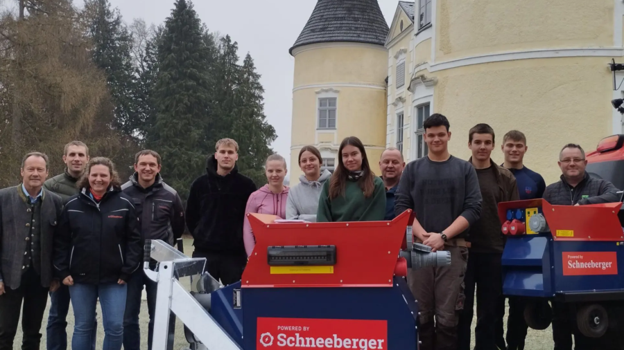 Gruppenfoto im Freien: Personen stehen hinter einem rot-blauen Schneeberger Gerät vor einem Gebäude mit Turm.