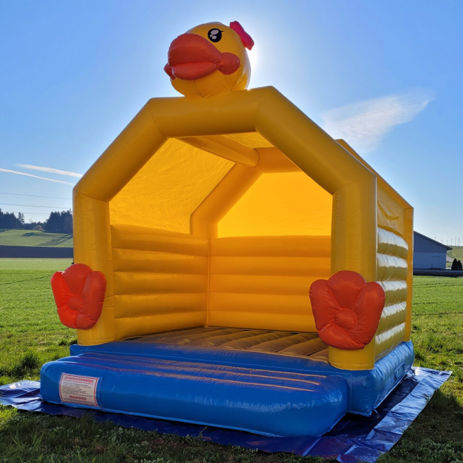 Gelbe Hüpfburg in Form eines Hühnerstalls steht im Freien auf einer Wiese.