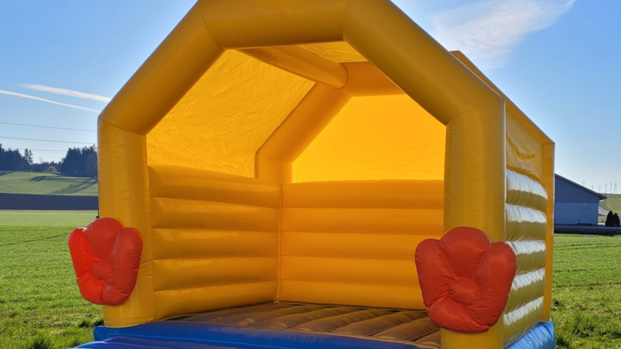 Gelbe Hüpfburg in Form eines Hühnerstalls steht im Freien auf einer Wiese.