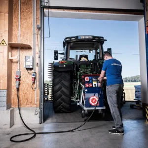 Traktor mit Zapfwellengenerator steht im Hallentor, ein Mitarbeiter bedient das Aggregat, Kabel führt zur Hausinstallation.