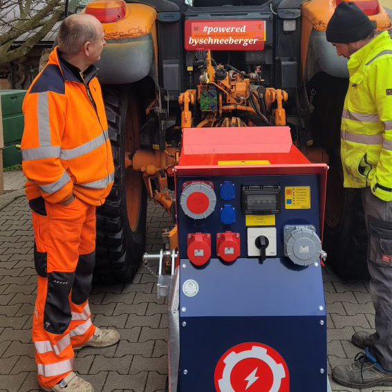 Zwei Personen in Arbeitskleidung stehen neben einem Traktor; davor steht ein rot-blauer Schneeberger Stromverteiler mit Steckdosenfeld.
